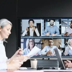 Mujer sentada en una mesa con un gran monitor a su izquierda. Videollamada con varios invitados.