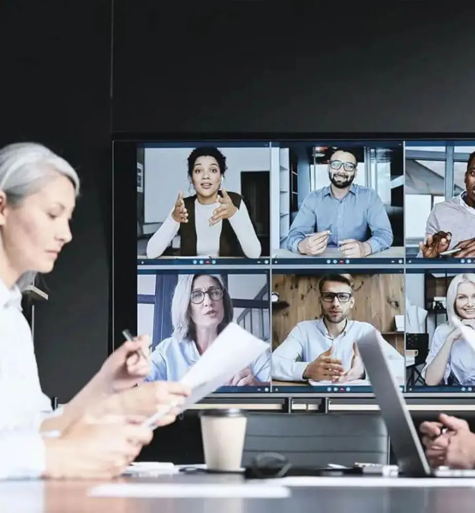 Mujer sentada en una mesa con un gran monitor a su izquierda. Videollamada con varios invitados.