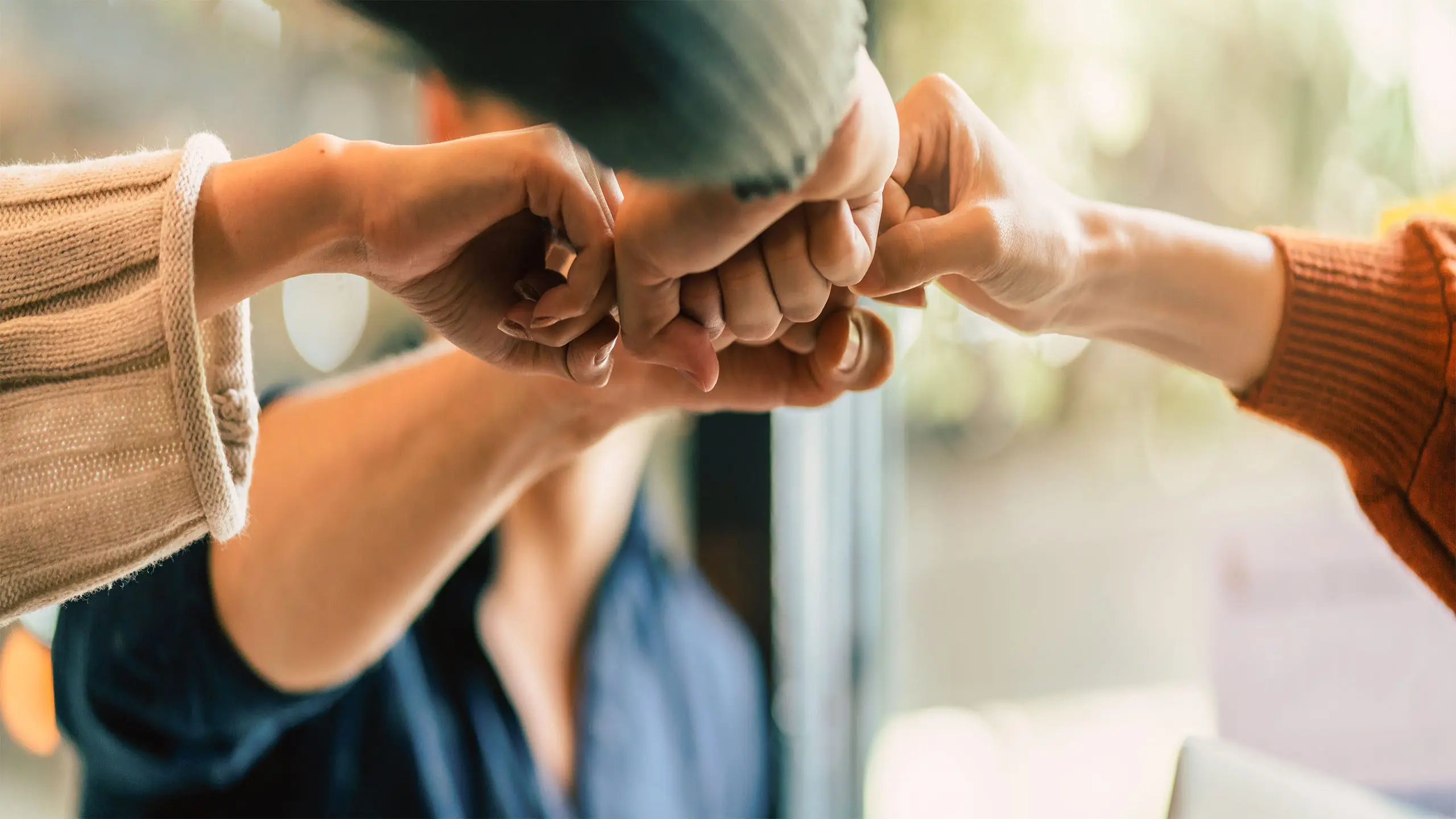 Six adults join their fists in solidarity at chest level. The image is zoomed in on just the hands.