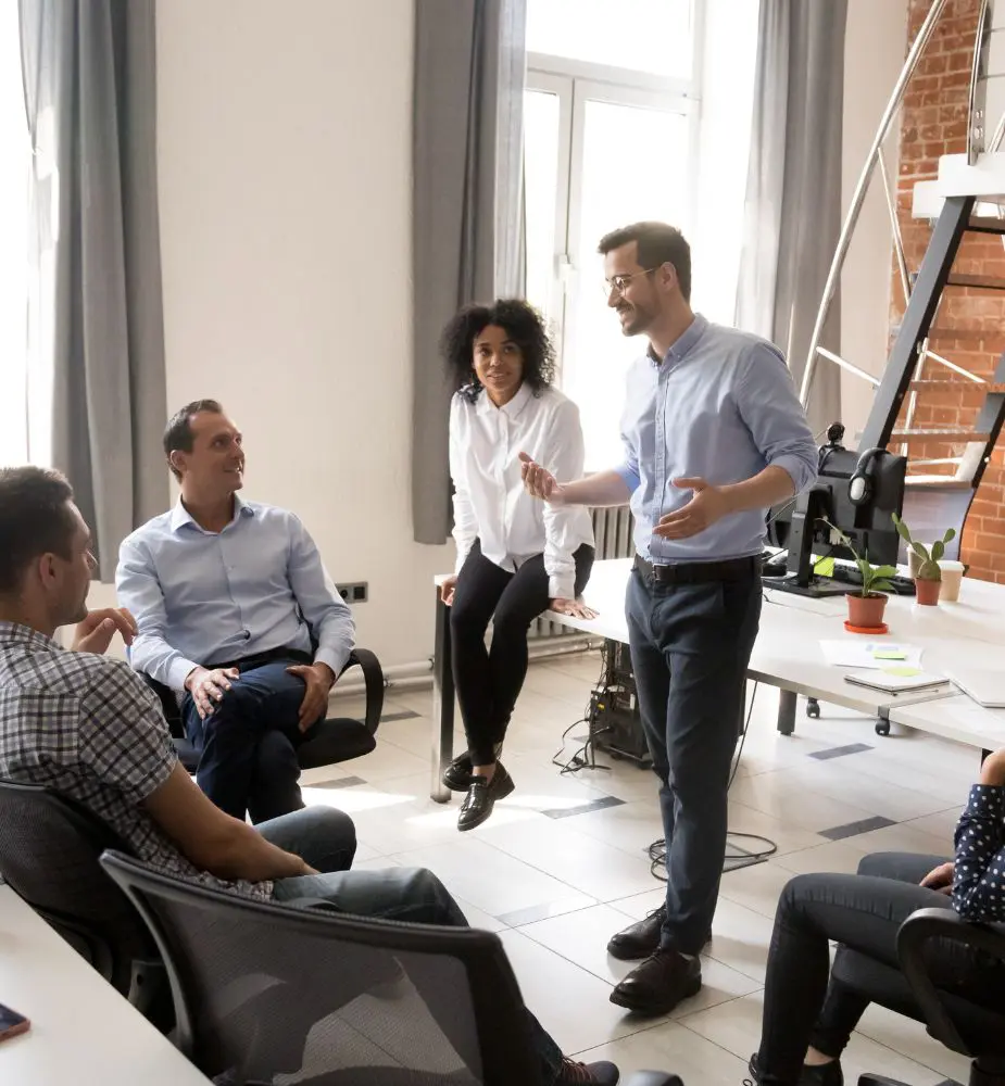 A leader stands and speaks casually to a small group seated in office chairs and one on a table.