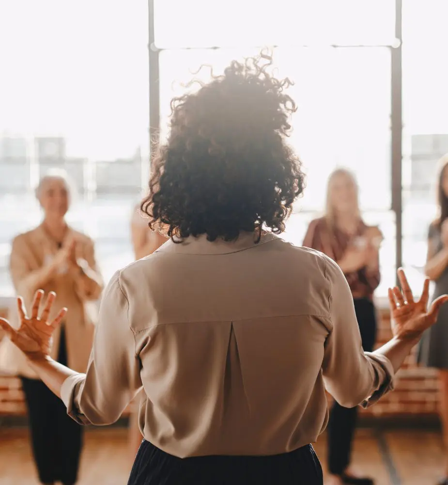 A woman faces a group of standing coworkers. Her back is to the camera and her hands are out as she presents an idea to the group.