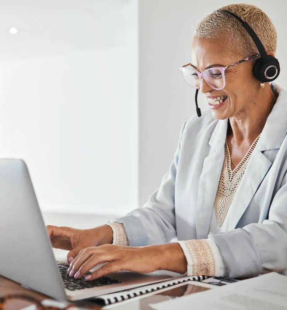 A woman in a headset sits at her laptop working and smiling. She looks to be on a call.