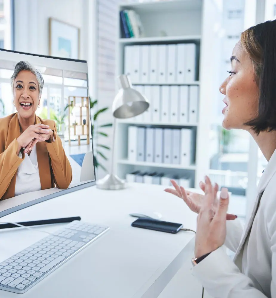 A woman in white faces her monitor as she has a video call with another woman in a mustard colored cardigan.