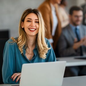 Una mujer con blusa azul está sentada detrás de su ordenador portátil sonriendo a la cámara. Hay compañeros de trabajo en una mesa detrás de ella.