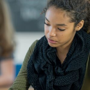 Student at a desk writing with a pencil. Another student is blurry in the background.